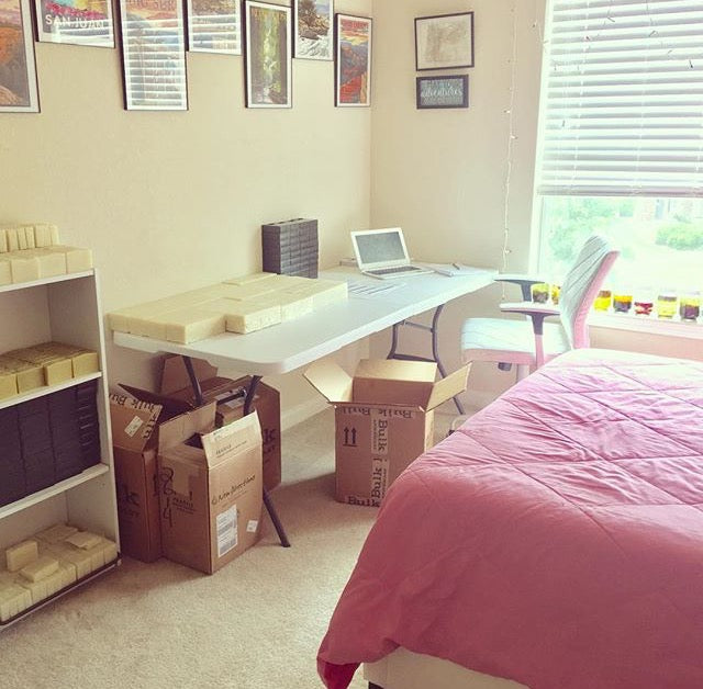 A room with soap bars on a shelf, a bed, a desk and boxes of packaged product.