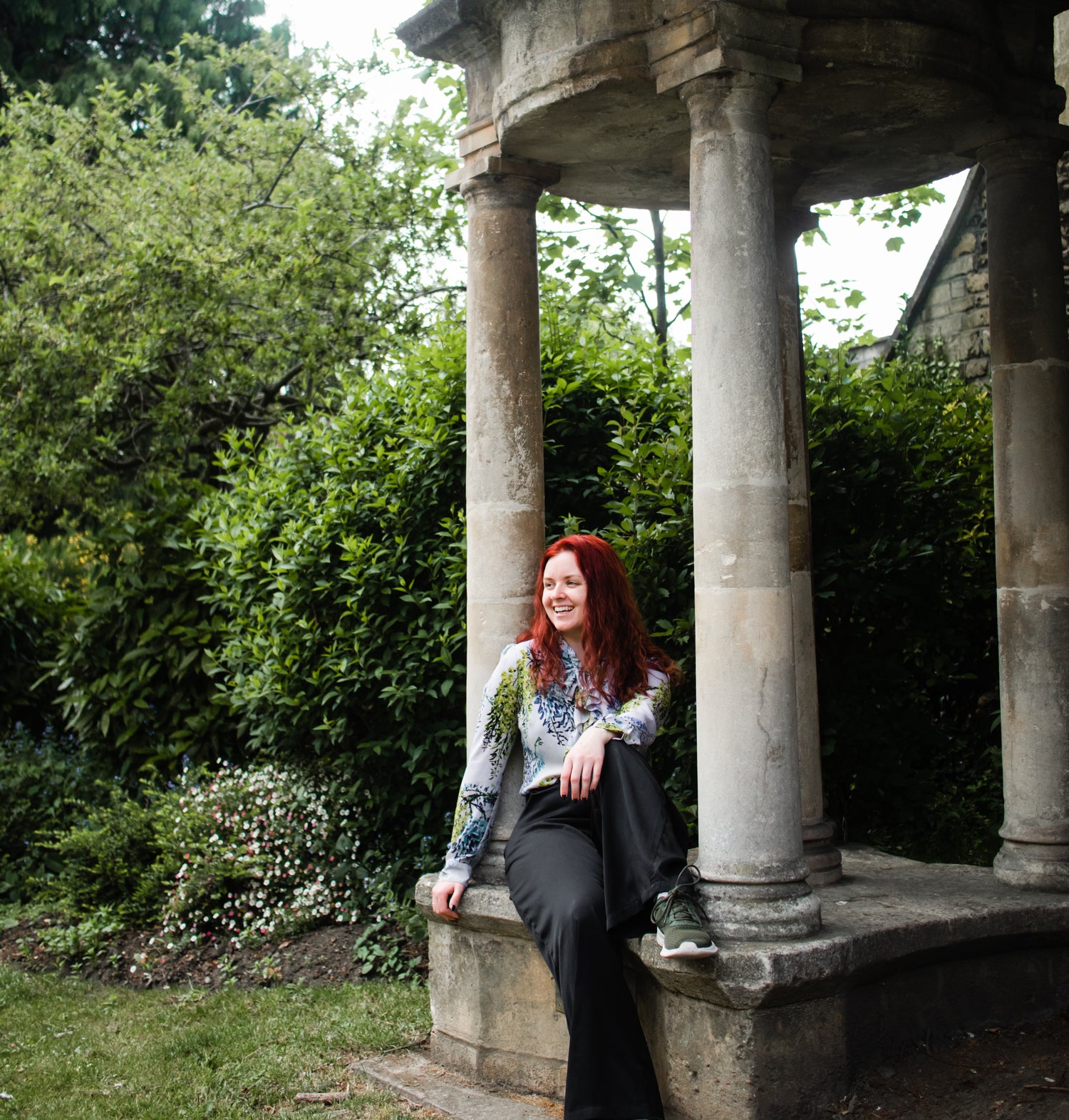 A woman sitting in front of green foliage on a stone column structure.  