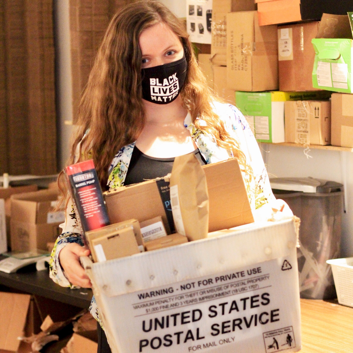 A young woman holding an United States postal office tub of packages while wearing a black lives matter mask.