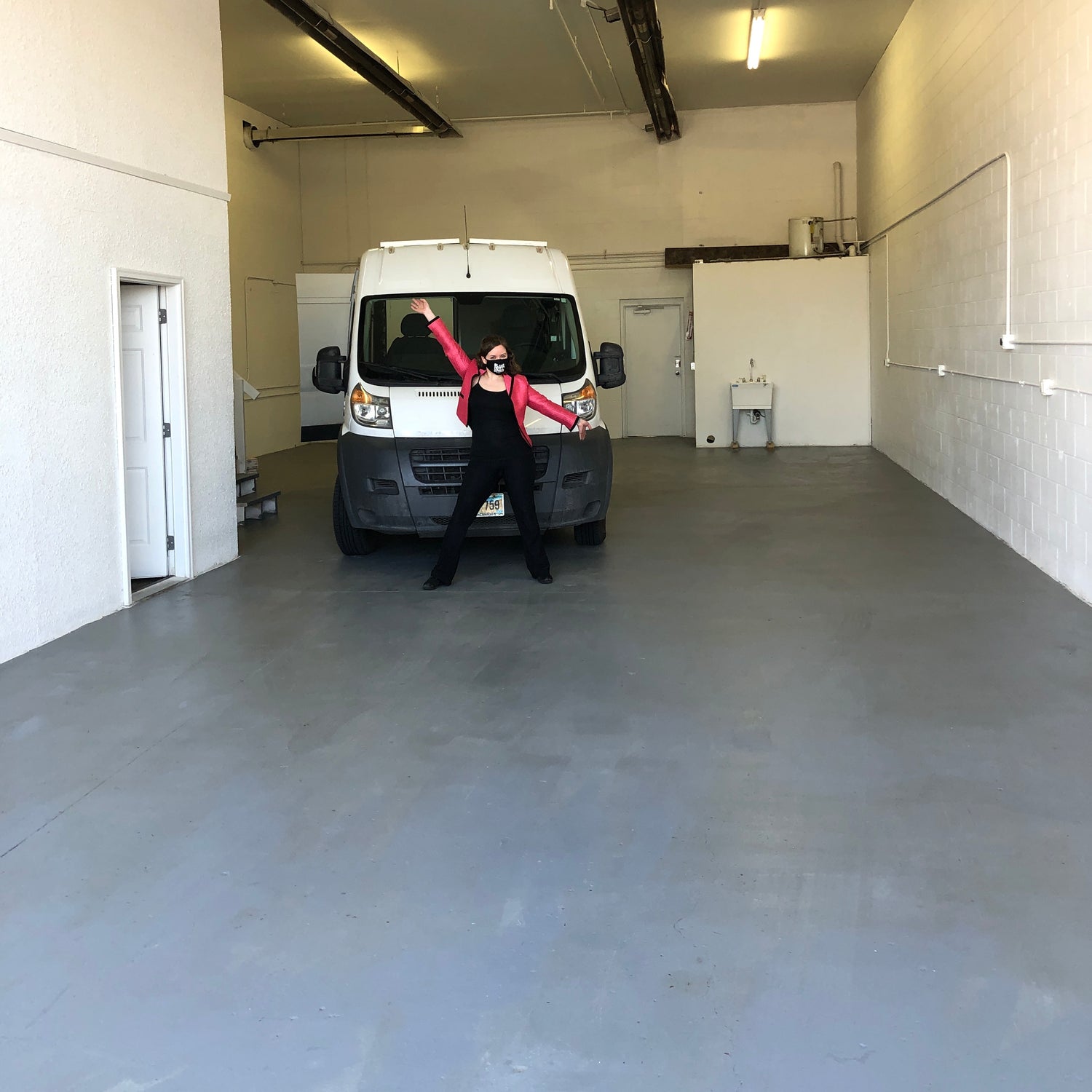 A young woman in an all black outfit and a pink blazer standing in front of a white cargo van in a warehouse with her arms up.