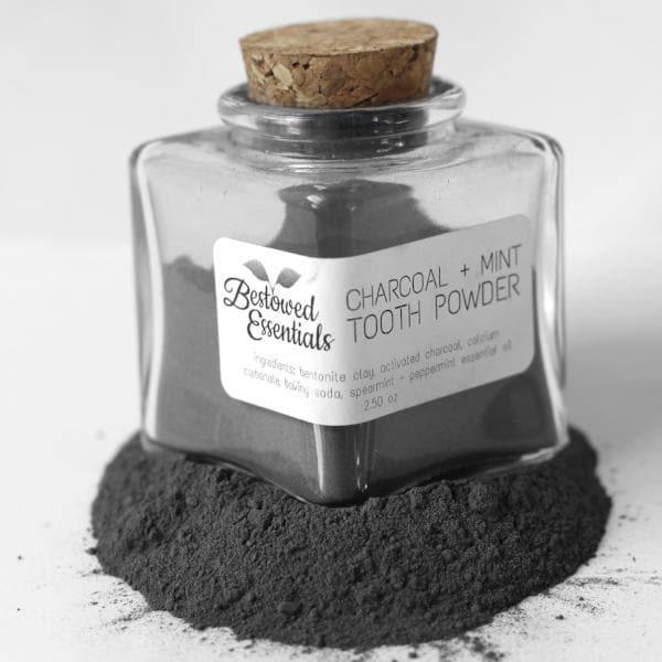 A square glass bottle with a cork top of gray tooth powder, sitting on the same gray powder with a white background.
