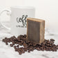Brown soap bar with black coffee specks sitting on coffee beans with a coffee mug behind it.