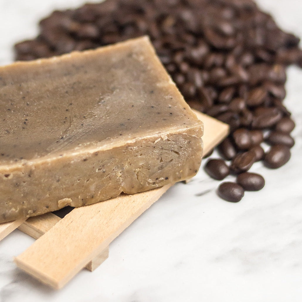 Brown soap bar with black coffee specks on a wooden soap holder with coffee beans splayed out next to the soap.