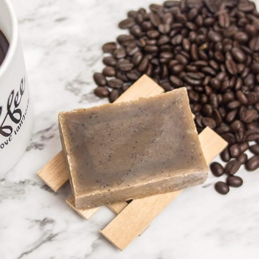 Brown soap bar with black specks of coffee sitting on top a wooden soap holder, with coffee beans splayed out next to it on a marble counter top.