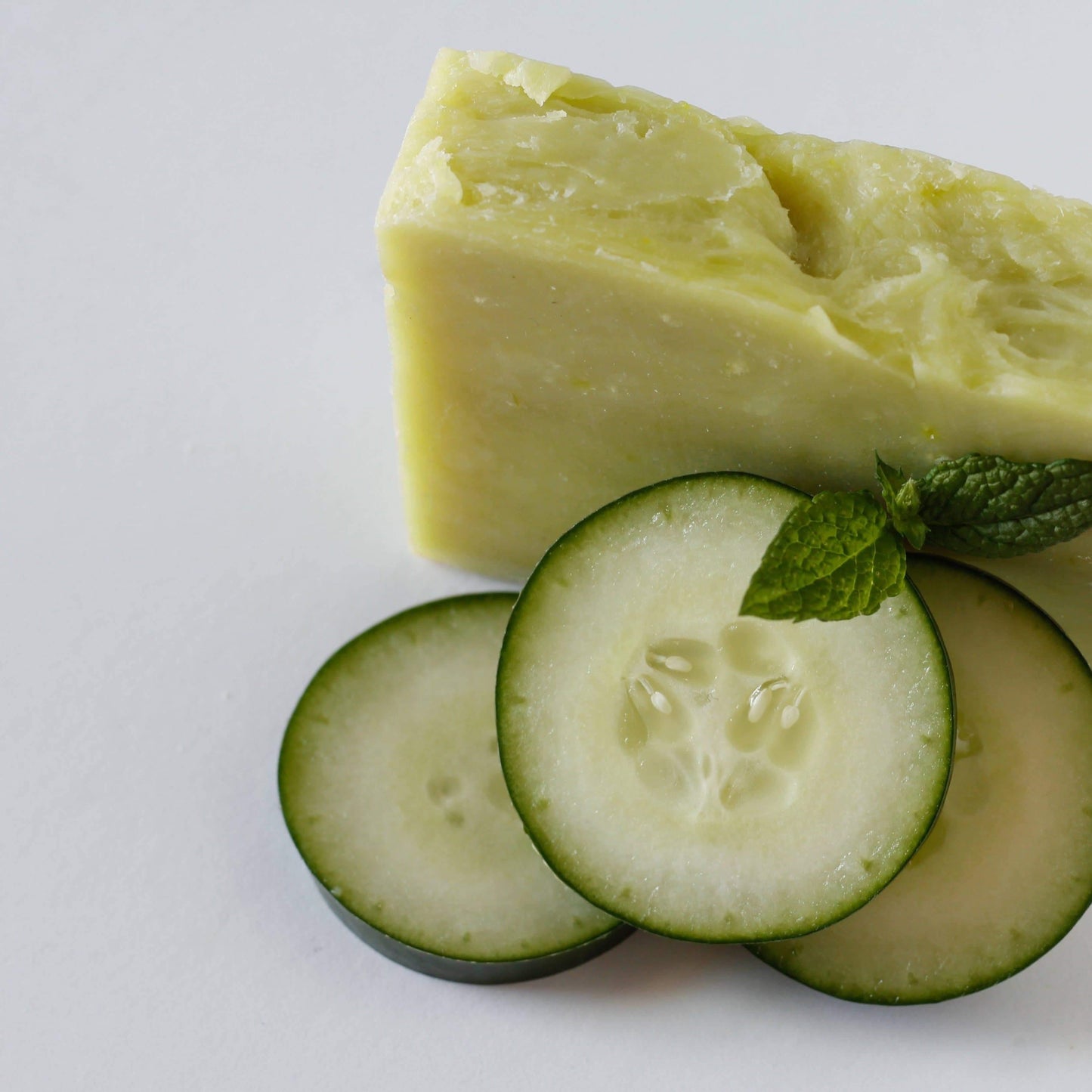 Light green soap bar on a white background decorated with cucumber slices and spearmint leaves.