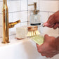 Hands scrubbing a bowl over the sink with a scrub brush. Tiled background with a bar of soap and gold faucet.