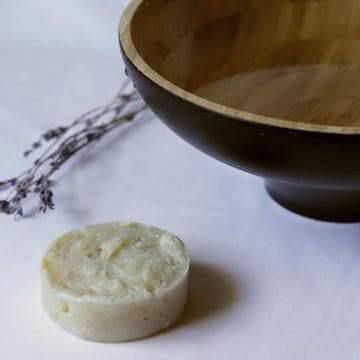 Circular shampoo bar next to a wooden bowl of water and a stalk of lavender on a white background.