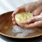 Cream colored shampoo bar being lathered in hands over a wooden bowl of soapy water.