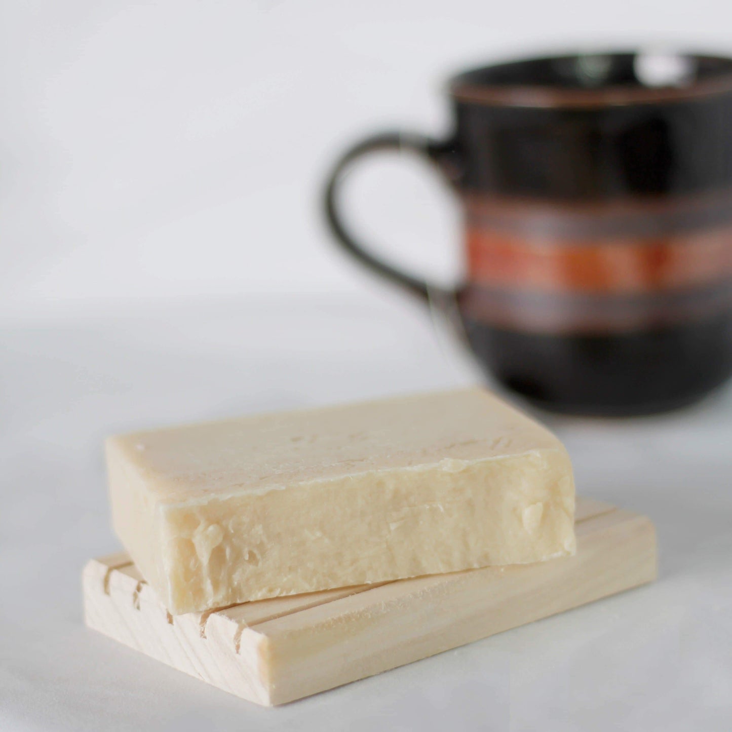 Cream colored soap bar on a wooden soap holder with a mug in the background.