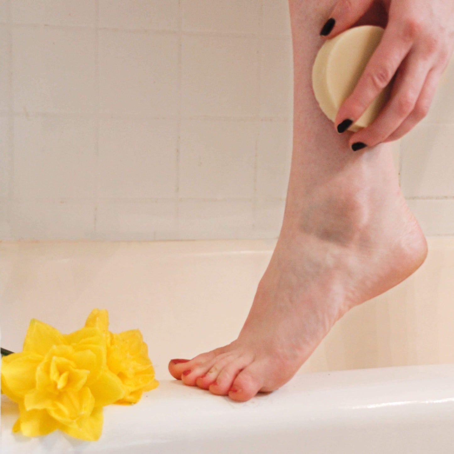 A woman rubbing a circular lotion bar on her leg over a bathtub with a bright yellow flower sitting on the tub.