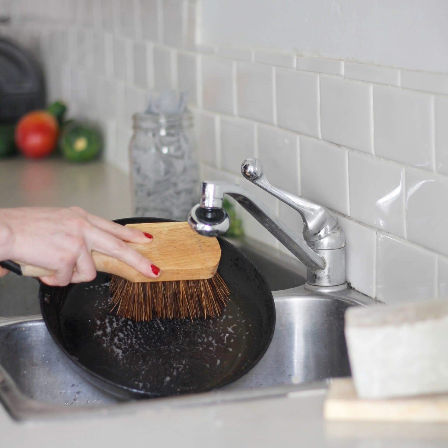 A woman scrubbing a pan over a sink with a wooden scrub brush with a white tiled background.