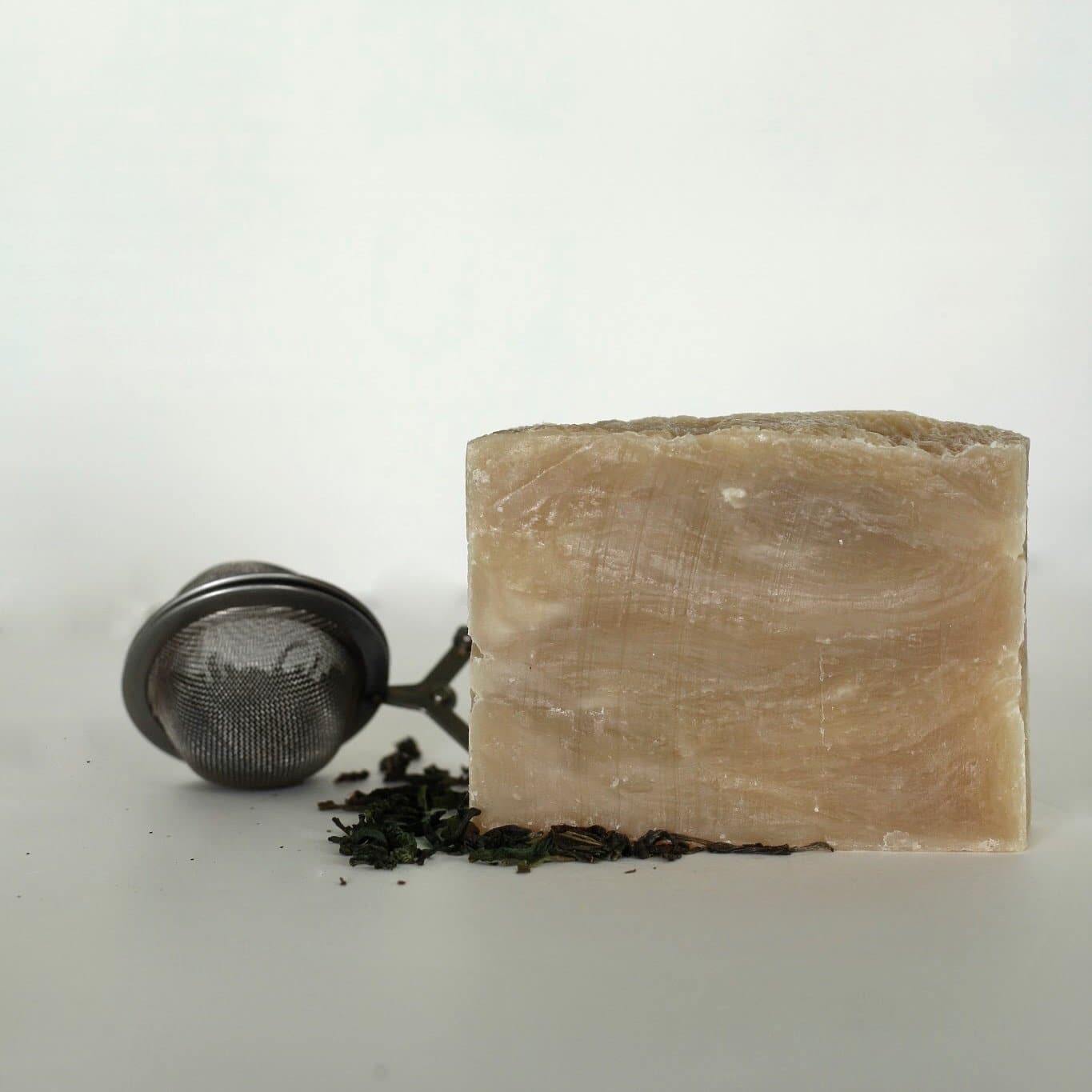 Cream soap bar sitting on top green tea leaves with a metal tea strainer next to it.